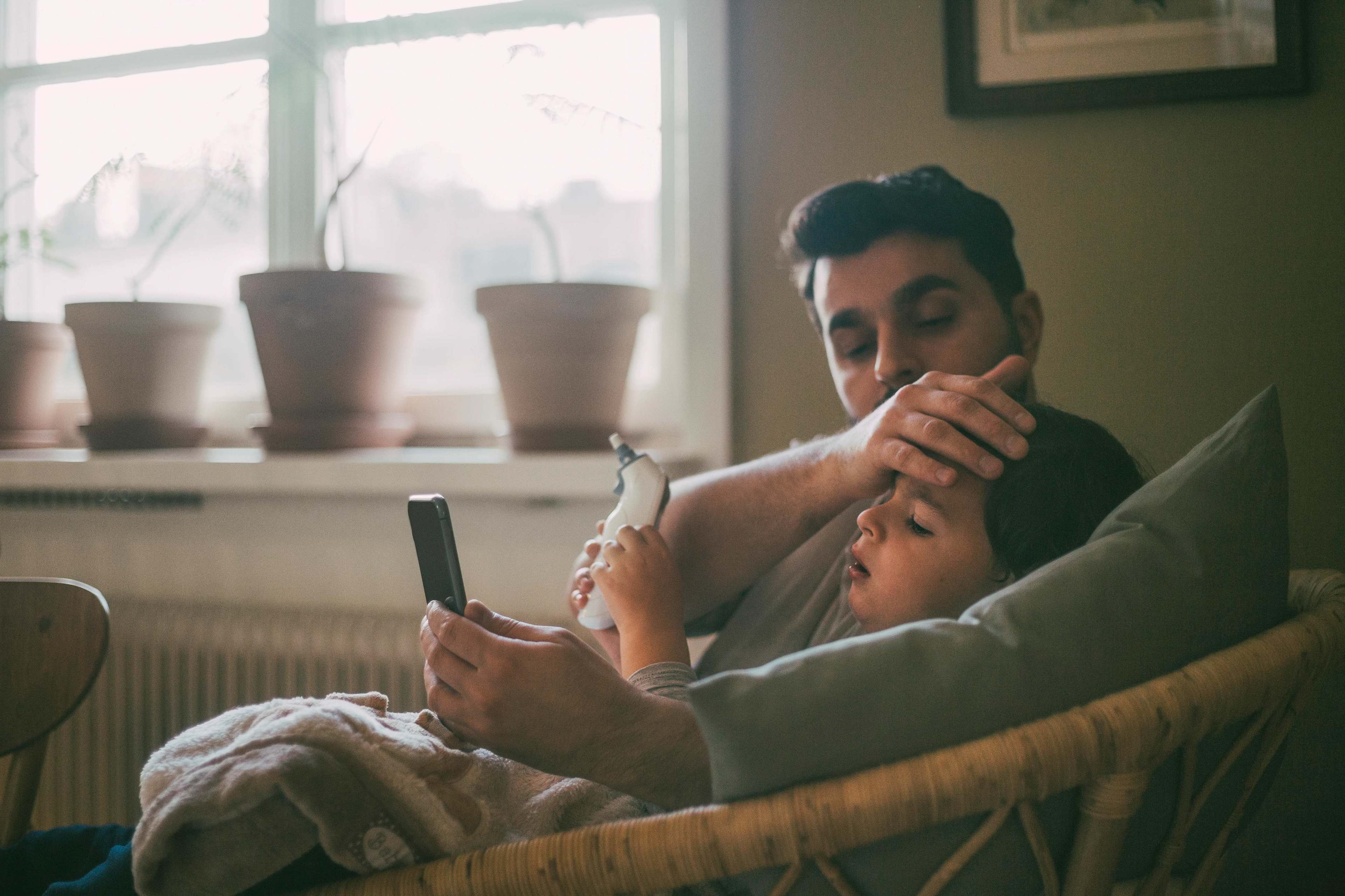 Gesundheit_Zuhause_Vater_Kind_Krank_Fiebermesser_Smartphone_s_eyimtt